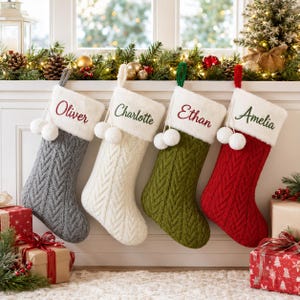 May include: Four personalized Christmas stockings featuring a cable knit pattern in gray, white, olive green, and red. Each stocking has a white faux fur cuff with two hanging pom-poms and embroidered names: Oliver, Charlotte, Ethan, and Amelia.