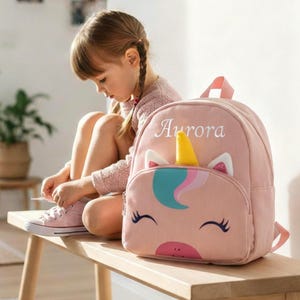 Pode incluir: Mochila rosa com design de rosto de unicórnio, com chifre amarelo, crina azul e rosa e o nome "Aurora" bordado na frente. A mochila tem um pequeno bolso frontal e alças rosa.