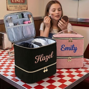 Può includere: Borse per il trucco in velluto a coste nere e rosa con cerniere dorate. La borsa nera ha "Hazel" ricamato in rosa, e la borsa rosa ha "Emily" ricamato in blu. La borsa nera aperta mostra pennelli e strumenti per il trucco.