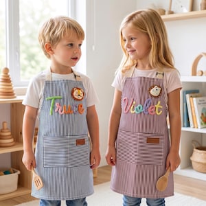 May include: Two children wearing striped aprons with personalized names and animal patches. One apron is blue and white, the other is burgundy and white. Each apron has a pocket and a wooden spoon.