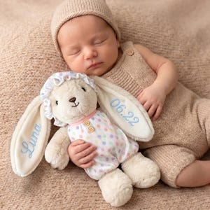 May include: A plush cream-colored rabbit toy wearing a white bonnet with colorful polka dots and a matching romper with two small wooden buttons. The rabbit's long ears are embroidered with the name Luna and the date 06.22 in light blue thread.