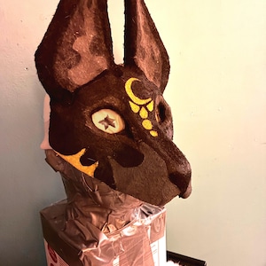 May include: A dark brown animal mask with tall, pointed ears and a textured surface. The mask features a yellow crescent moon and dot design on the forehead, and a star-shaped pattern within the eye socket area, mounted on a box base.