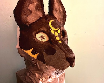 May include: A dark brown animal mask with tall, pointed ears and a textured surface. The mask features a yellow crescent moon and dot design on the forehead, and a star-shaped pattern within the eye socket area, mounted on a box base.