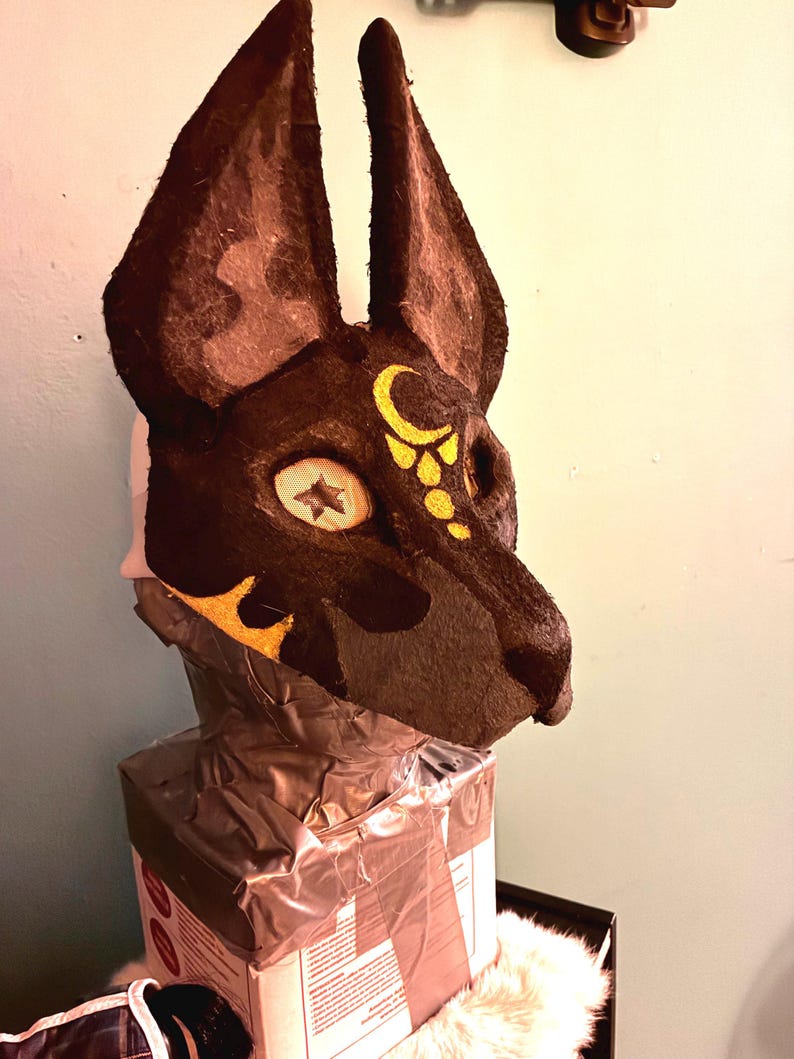 May include: A dark brown animal mask with tall, pointed ears and a textured surface. The mask features a yellow crescent moon and dot design on the forehead, and a star-shaped pattern within the eye socket area, mounted on a box base.