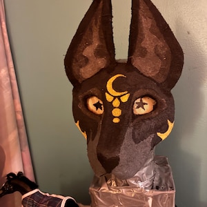 May include: A black and dark gray animal mask featuring tall, pointed ears and a gold crescent moon symbol on the forehead. The mask has mesh eye coverings with black star details and gold accents on the cheeks, mounted on a rectangular base.