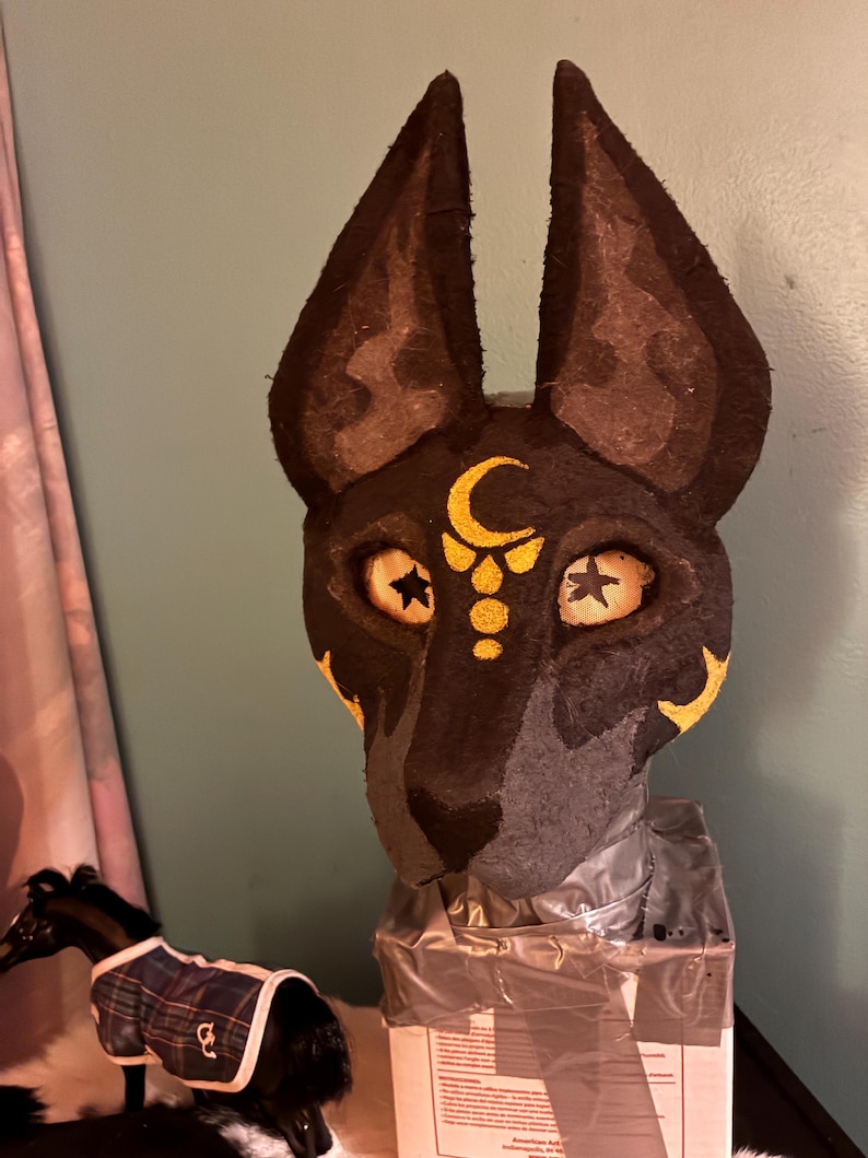 May include: A black and dark gray animal mask featuring tall, pointed ears and a gold crescent moon symbol on the forehead. The mask has mesh eye coverings with black star details and gold accents on the cheeks, mounted on a rectangular base.