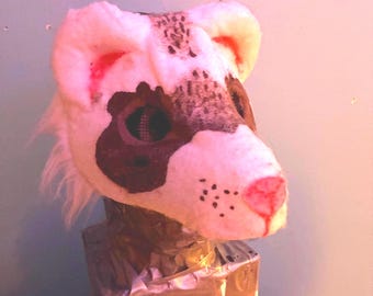 May include: A white and brown faux fur animal mask featuring pointed ears, dark eye patches, a pink nose, and black whisker spots. The mask is mounted on a rectangular base wrapped in silver duct tape, showcasing a textured, plush appearance.