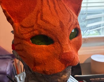 May include: A handmade orange cat mask with dark brown stripes. The mask has green eye lenses and is mounted on a mannequin head. The mask is made of a felt-like material.