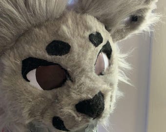 May include: A gray furry animal mask with black spots and large ears. The eyes are white with a brown mesh. The mask is attached to a cardboard box with gray tape. Blue lights are attached to the top of the mask.
