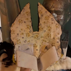 May include: A mask with tall, pointed ears featuring a cream-colored fabric lining with a small yellow and blue floral pattern. The interior includes a white elastic strap for securing the mask and a label reading The Dorset Collection.