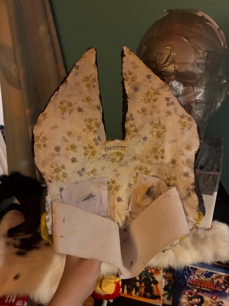 May include: A mask with tall, pointed ears featuring a cream-colored fabric lining with a small yellow and blue floral pattern. The interior includes a white elastic strap for securing the mask and a label reading The Dorset Collection.