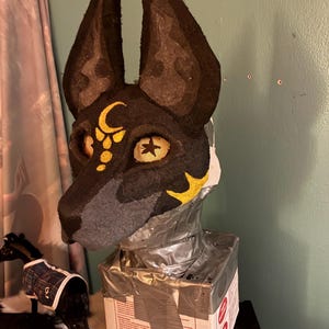 May include: A black and grey animal mask featuring tall, pointed ears, yellow eye sockets with star-shaped pupils, and a gold crescent moon symbol on the forehead. The mask is mounted on a rectangular base wrapped in silver duct tape.