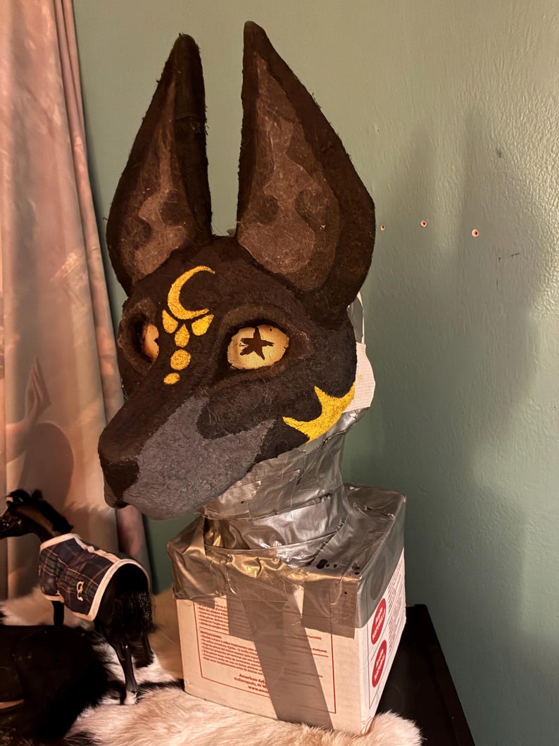 May include: A black and grey animal mask featuring tall, pointed ears, yellow eye sockets with star-shaped pupils, and a gold crescent moon symbol on the forehead. The mask is mounted on a rectangular base wrapped in silver duct tape.