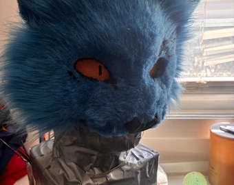 May include: A blue furry cat mask with orange eyes and a black nose. The mask is mounted on a box covered in gray tape. The mask has a realistic design, with detailed fur and facial features. The mask is likely for cosplay or costume use.