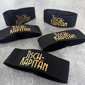 Könnte beinhalten: Fünf schwarze elastische Armbänder mit dem goldfarbenen Aufdruck "Tisch-Kapitän". Die Armbänder sind auf einer grauen Oberfläche angeordnet. Der Text ist in einer stilisierten Schriftart.