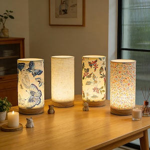 May include: Four cylindrical table lamps with wooden bases and fabric shades. The shades display diverse designs, including butterflies, floral patterns, and a textured neutral fabric. The lamps are illuminated, emitting a warm glow, and are placed on a wooden table.