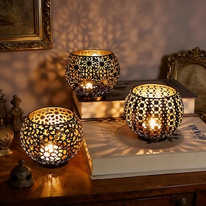 Peut inclure: Trois bougeoirs sphériques en métal noir avec des motifs ajourés complexes. Chaque bougeoir contient une bougie allumée, projetant une lumière chaude et des ombres décoratives. Les bougeoirs sont posés sur des livres.