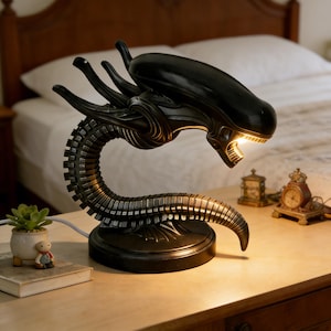 Op de afbeelding: Een zwarte Alien-thema lamp met een gedetailleerd, gestructureerd ontwerp. De lamp heeft een Xenomorph-hoofd met een open mond, die een warme gloed uitstraalt. De nek van de lamp buigt in een voet, en staat op een houten oppervlak.