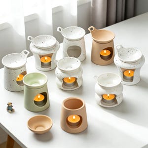 May include: A collection of ceramic oil warmers in various shapes and colors, including white, beige, and green. Each warmer has a small dish on top and a space for a lit tea light candle, creating a warm, inviting ambiance.