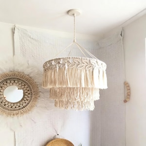 Op de afbeelding: Een crèmekleurige macramé kroonluchter met drie niveaus hangt aan het plafond. De kroonluchter heeft een cirkelvormig ontwerp met ingewikkeld knoopwerk en lange franjes. De lamp wordt opgehangen door een gevlochten koord, wat een bohemien touch aan de kamer geeft.
