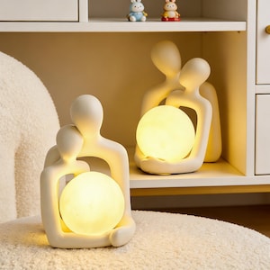 Może przedstawiać: Biały ceramiczny kubek z czarnym uchwytem i napisem "But First, Coffee" pogrubioną, czarną czcionką. Kubek jest umieszczony na rozmytym tle, sugerującym przytulne otoczenie.