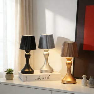 Può includere: Tre lampade da tavolo decorative con paralumi plissettati neri, argentati e dorati. Ogni lampada ha una base curva e poggia su una superficie bianca. Le lampade sono disposte in fila, mostrando i loro diversi colori e design.