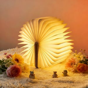 May include: A unique, open book-shaped lamp with warm, glowing pages. The lamp is made of light-colored material and has a dark brown spine. The lamp is surrounded by flowers and small figurines, creating a cozy and inviting atmosphere.