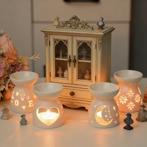 Op de afbeelding: Vier crèmekleurige keramische oliebranders met uitgesneden ontwerpen van harten, sterren en letters. Een kleine kast met glazen deuren en beeldjes staat op de achtergrond. Elke brander heeft een brandende kaars.