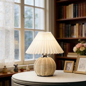 Pode incluir: Uma luminária de mesa creme com base em vime tecido e abajur plissado. A luminária está sobre uma mesa redonda e branca. O fundo inclui uma janela com cortina de renda, uma estante e fotos emolduradas.