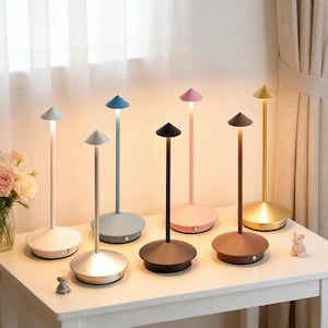 May include: A collection of modern, minimalist table lamps in various colors, including white, blue, pink, brown, and gold. Each lamp features a conical shade and a slender stem, with a circular base. The lamps are arranged on a white table.