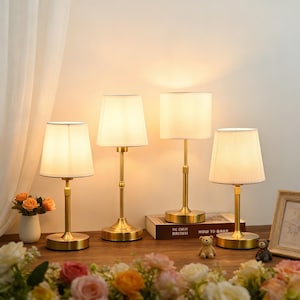 Puede incluir: Cuatro lámparas de mesa doradas con pantallas blancas. Las lámparas están dispuestas sobre una superficie de madera. Las pantallas son de color crema y las lámparas están encendidas, emitiendo un brillo cálido. Las lámparas miden unos 30 cm de alto.