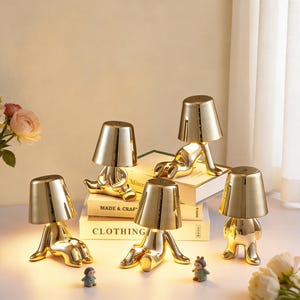 May include: A collection of gold-colored table lamps in a whimsical human-like design. Each lamp has a conical shade and a body shaped like a person in various poses. Some lamps are placed on books with text that reads "MADE & CRAFT CLOTHING".