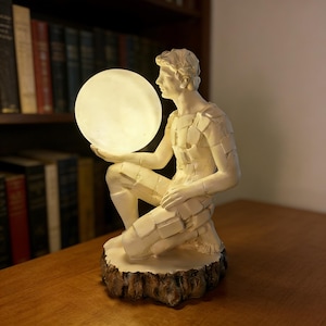 Puede incluir: Lámpara decorativa de color crema que representa una figura arrodillada sosteniendo un orbe brillante. La figura está esculpida con un patrón geométrico texturizado en la ropa. La lámpara se asienta sobre una base marrón con forma de tocón de árbol.