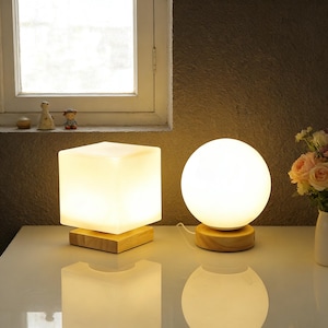 Pode incluir: Duas luminárias de mesa iluminadas. Uma tem formato de cubo branco, e a outra de esfera branca. Ambas as luminárias estão sobre uma base quadrada de madeira. As luminárias estão sobre uma superfície branca, com uma janela e flores ao fundo.