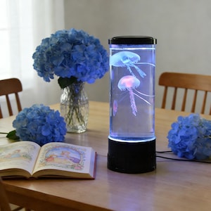 Puede incluir: Un acuario cilíndrico para medusas con base y tapa negras, iluminado con luces azules y rosas. Dos medusas flotan en el interior. Flores de hortensia azules en un jarrón y sobre la mesa completan la escena.