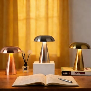 May include: Three modern mushroom-shaped lamps in copper, silver, and gold tones are displayed on a wooden surface. The lamps have a conical base and a rounded top, with a warm glow emanating from beneath. An open book and a pen are in the foreground.