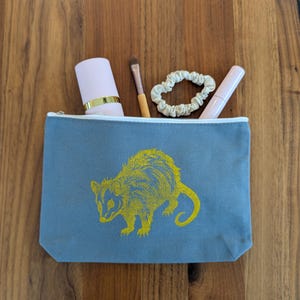 May include: A blue-grey zipped pouch featuring a yellow possum design. Inside the pouch are a pink lipstick, a makeup brush, a scrunchie, and a lip gloss. The pouch rests on a wooden surface.