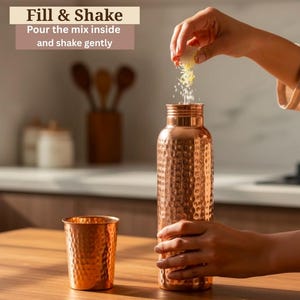 Könnte beinhalten: Eine Kupferwasserflasche und ein passender Becher mit gehammerter Textur. Die Flasche wird mit einer Mischung gefüllt. Der Text auf dem Bild lautet: "Füllen & Schütteln. Die Mischung hineingießen und vorsichtig schütteln."