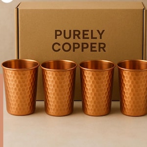 May include: Four copper-colored drinking cups with a hammered texture are displayed in front of a cardboard box. The box has the words "PURELY COPPER" printed on it. The cups are cylindrical and have a shiny finish.