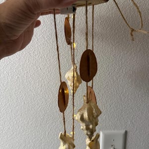 Peut inclure: Un mobile suspendu composé d'une base en bois circulaire surmontée de verre de mer jaune et éclairée par de petites lumières LED. Des brins de ficelle pendent de la base, ornés de disques bruns plats et de coquillages blancs en couches.