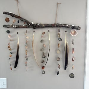 Könnte beinhalten: Ein Wandbehang bestehend aus einem horizontalen Holzast, der an einer Schnur aufgehängt ist. An dem Ast sind mehrere vertikale Stränge aus Muscheln und langen, dunkel gefiederten Federn befestigt. Die Muscheln variieren in Größe, Form und Farbe, einschließlich Braun-, Weiß- und Grautönen, und bilden eine kaskadierende Anordnung.