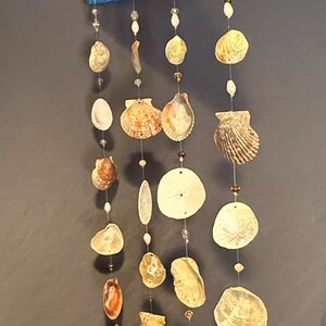 May include: A coastal-themed mobile featuring a painted wooden top in blue, yellow, and white hues, resembling a sunset. Various seashells and beads are strung below, creating a decorative hanging ornament.