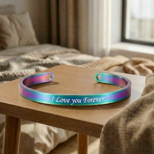 Peut inclure: Un bracelet manchette aux couleurs de l'arc-en-ciel avec l'inscription "I Love you Forever" en lettres blanches. Le bracelet est en métal et a un design incurvé. Le bracelet est présenté sur une table en bois.
