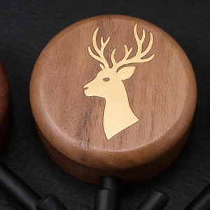 Op de afbeelding: Houten flessenstoppers met een gouden hertkopontwerp. De stoppers zijn gemaakt van donkerbruin hout en hebben een ronde vorm. De hertenkop is een silhouet met geweien. Op een zwarte achtergrond.