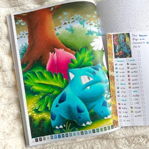 May include: A vibrant, colored illustration of a Bulbasaur Pok&eacute;mon in a forest setting, with a large tree and pink flowers. The image is from a coloring book, with a color key and the text "Tome Pokemon" visible.