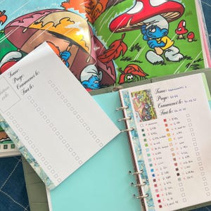 May include: An open binder with a teal cover and lined pages, featuring a colorful illustration of Smurfs. A notepad with a dotted grid and the text "Tome: Pages Commenc&eacute; le: Fini le:" is in the foreground. The background shows a Smurfs book.