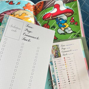 May include: A white Solution colo checklist with a dotted grid, and the words "Tome", "Page", "Commenc&eacute; le:" and "Fini le:" printed in black. The checklist is in front of an open book with a Smurf illustration.