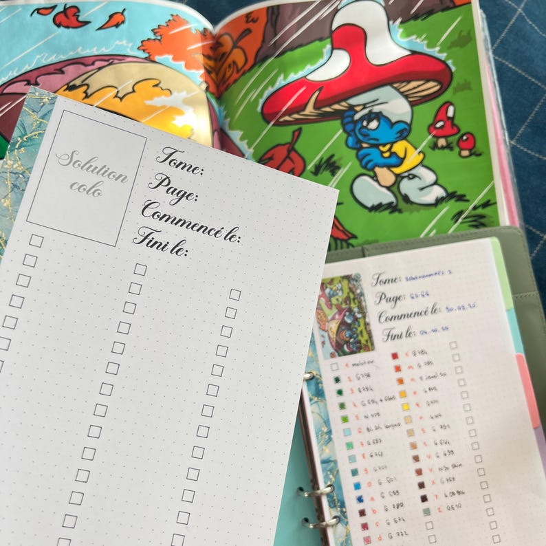 May include: A white Solution colo checklist with a dotted grid, and the words "Tome", "Page", "Commenc&eacute; le:" and "Fini le:" printed in black. The checklist is in front of an open book with a Smurf illustration.