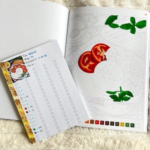 May include: Open coloring book page with a partially colored image of tomatoes, mozzarella, and basil. A small notepad with a checklist and text in French is on the left. The notepad is decorated with floral washi tape.