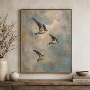 Puede incluir: Una obra de arte enmarcada que presenta tres golondrinas en vuelo contra un cielo nublado. Las aves tienen plumaje marrón y blanco con largas colas bifurcadas. El fondo es una mezcla de tonos azules, beige y blancos, creando un efecto suave y etéreo. El marco es marrón cálido.